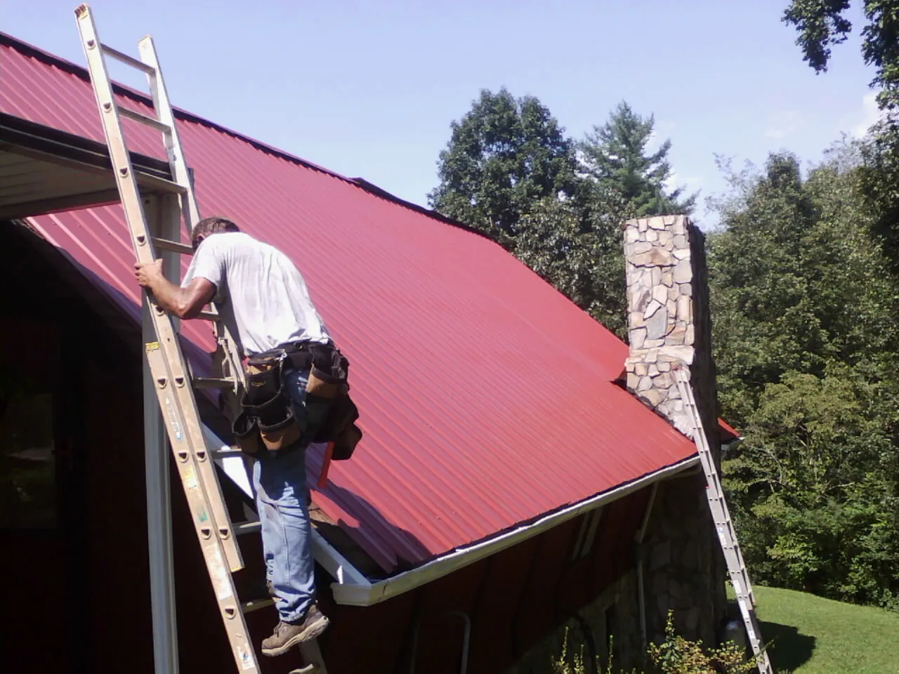 Roofer on a ladder beside a red metal roof and stone chimney — Soffit & Fascia Repair by Roofing Brothers in Star Junction